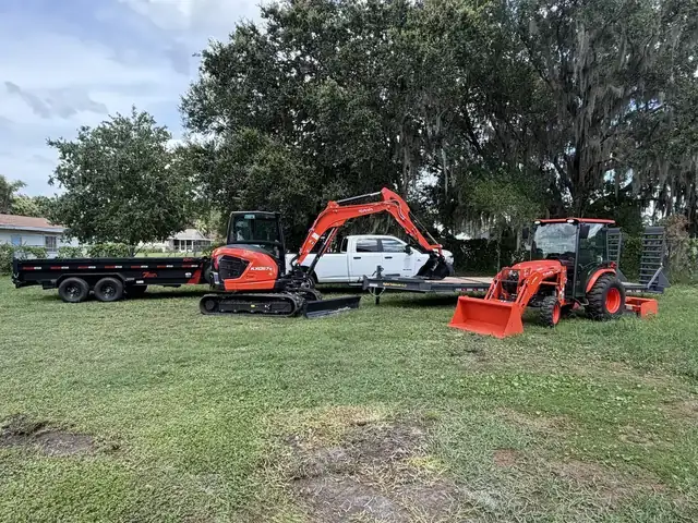 Faith Works Outdoor Services fleet — Kubota excavator, tractor with loader, pickup, dump trailer, and flatbed trailer